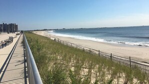 Ein Strand mit Promenadenweg, grasbewachsenen Dünen und blauem Himmel.