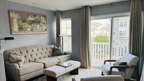 Ein Wohnzimmer mit einem Ecksofa, einer Chaiselongue und einem Couchtisch. Ein großes Fenster mit Vorhängen und ein Gemälde an der Wand.