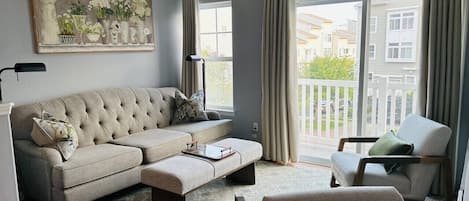 A living room with a sectional sofa, a chaise lounge, and a coffee table. There is a large window with curtains, and a painting on the wall.
