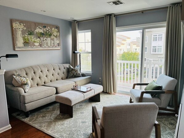 Una sala de estar con un sofá modular, una chaise longue y una mesa de centro. Hay un ventanal con cortinas y un cuadro en la pared.