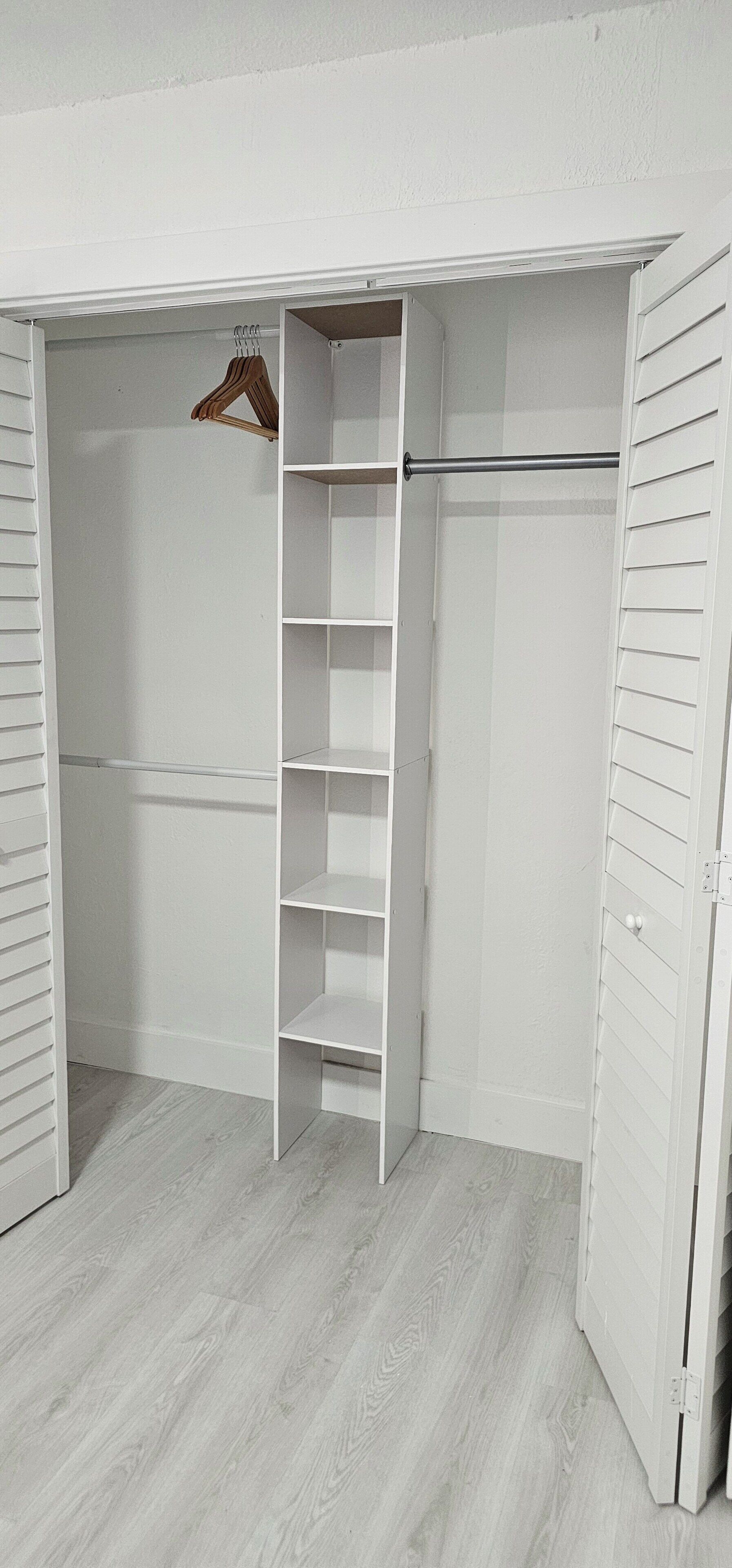 Un armario blanco con varias estanterías y una barra para colgar, suelo de madera y paredes blancas.