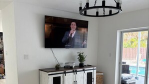 A living room with a flat-screen TV mounted on the wall, a white cabinet with black handles, and a large window with a view of a backyard.