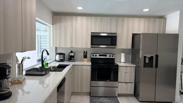 A modern kitchen with light wood cabinets, stainless steel appliances, a built-in oven, and a refrigerator.