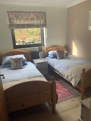Quarto com duas camas de madeira, uma mesa de cabeceira, janela com vista para pedras e vegetação, e cortina estampada.