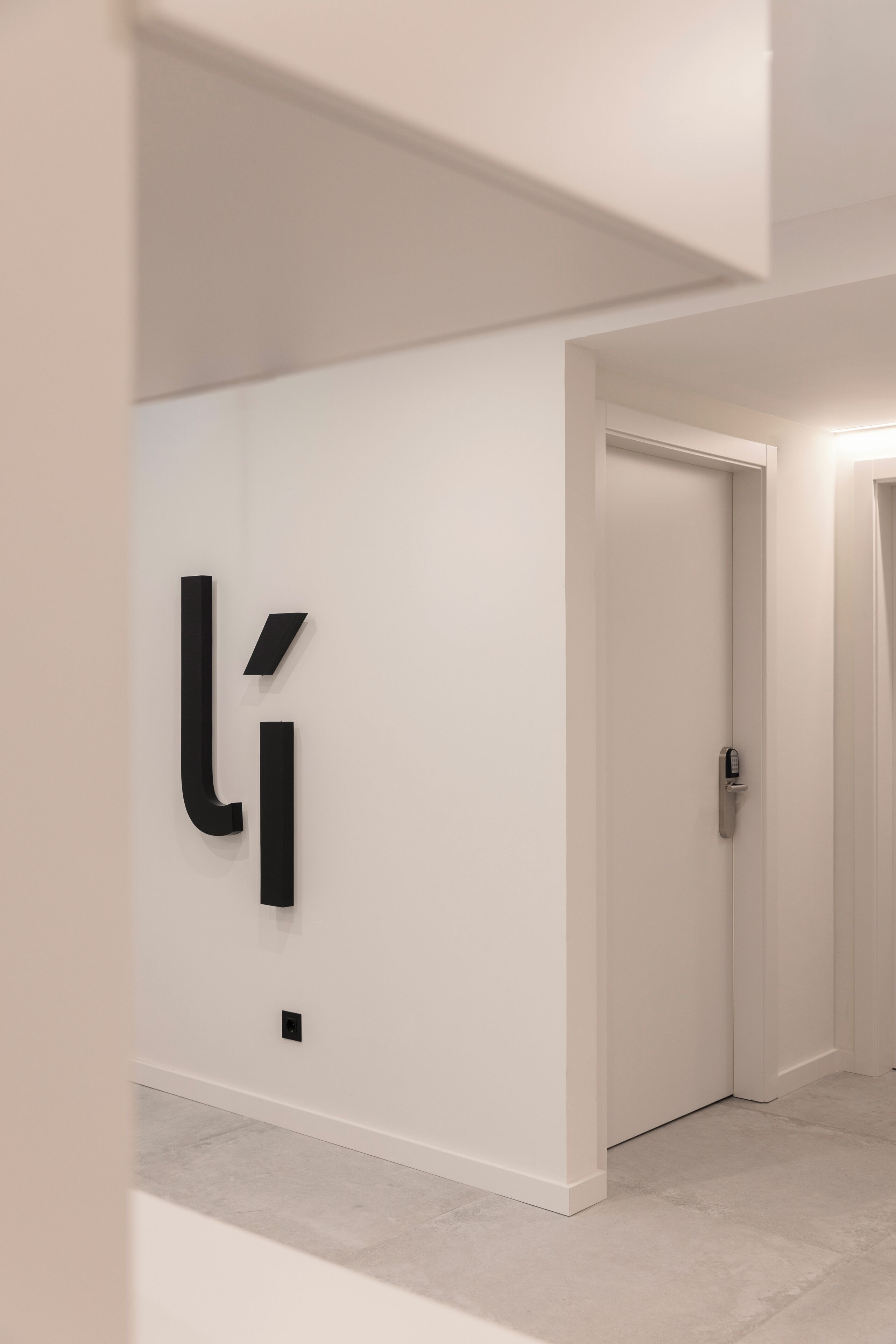 A hallway with a modern design, featuring a large black letter 'i' on the wall and a door with a keypad lock.
