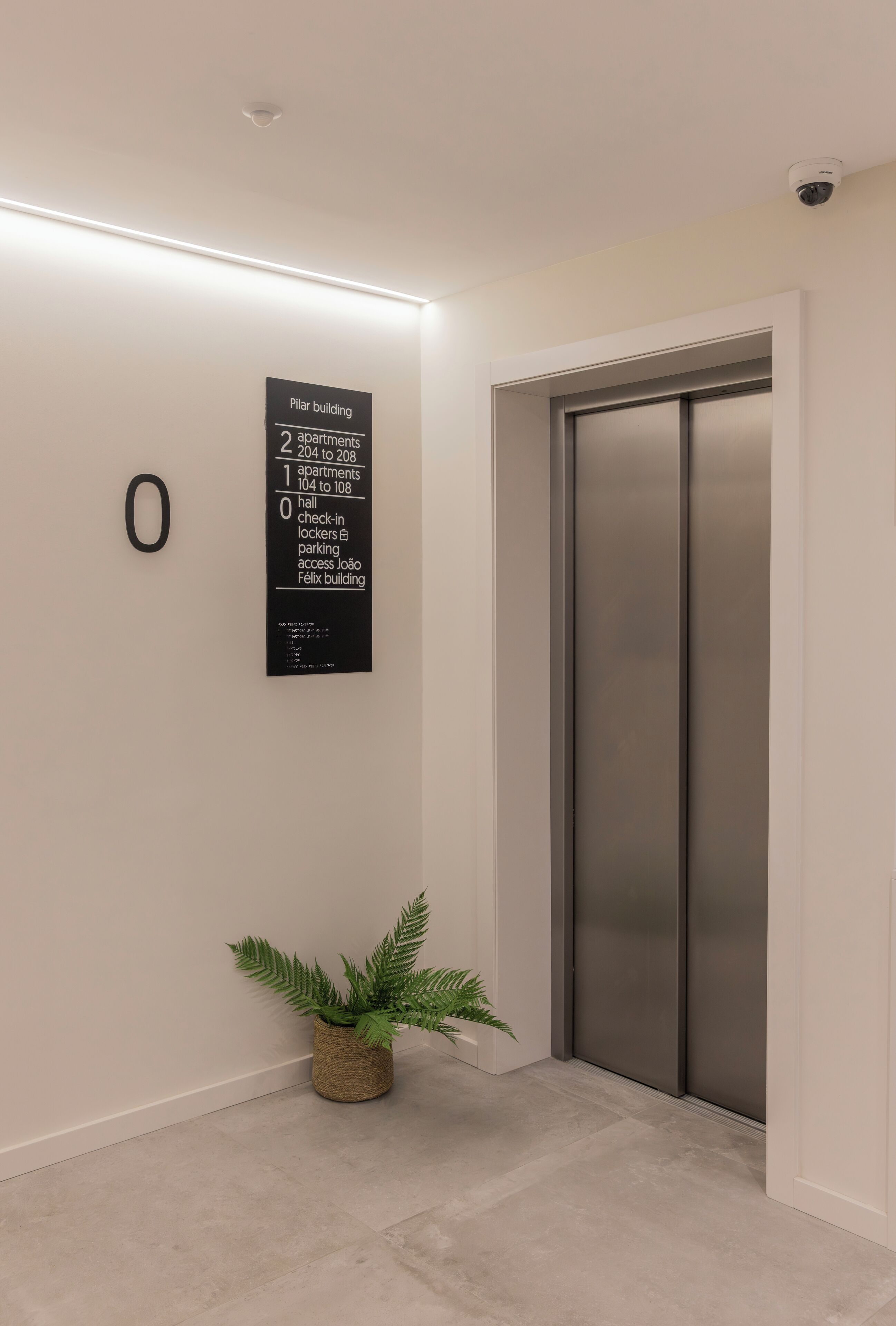 Ein moderner Aufzug mit digitaler Anzeige, einem Schild mit dem Namen des Gebäudes und Apartmentnummern sowie einer Zimmerpflanze im Eingangsbereich.