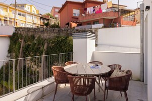 Un balcón con sillas de mimbre y una mesa, con vistas a una zona residencial con edificios y un muro de piedra.