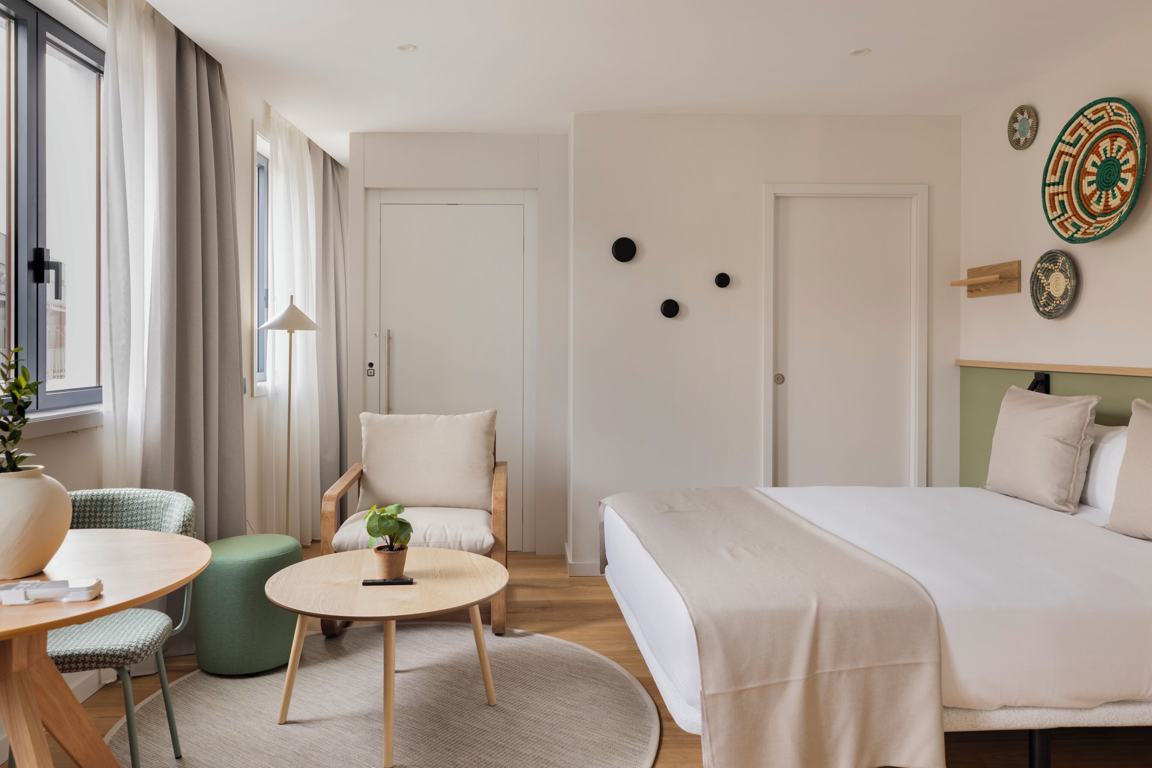A hotel room with a bed, a round table with a plant, a green ottoman, a chair, and a view of the city through the window.