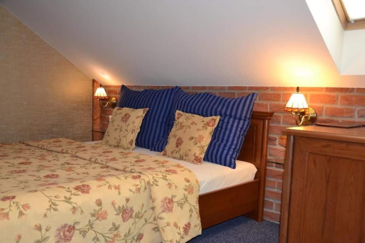 Ein Schlafzimmer mit schräg angesetzter Decke, einem hölzernen Bettgestell, einer blumigen Tagesdecke, gestreiften Kissen und zwei Wandleuchten.
