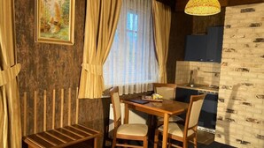 Une pièce de taille modeste, avec une table à manger en bois, quatre chaises, un petit meuble, une fenêtre avec des rideaux, un luminaire suspendu et un mur décoré d’un motif en briques.