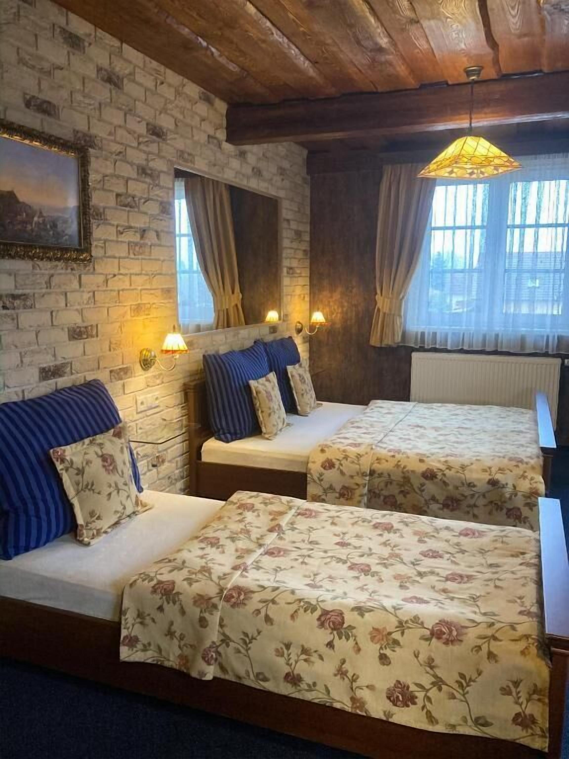 A room with two beds, a brick wall, wooden ceiling beams, and a window with curtains.