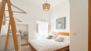 Ein Schlafzimmer mit einem hölzernen Bett, einem kleinen Nachttisch, einem Fenster mit Vorhängen, einer Pendelleuchte und einem an der Wand hängenden Bild.