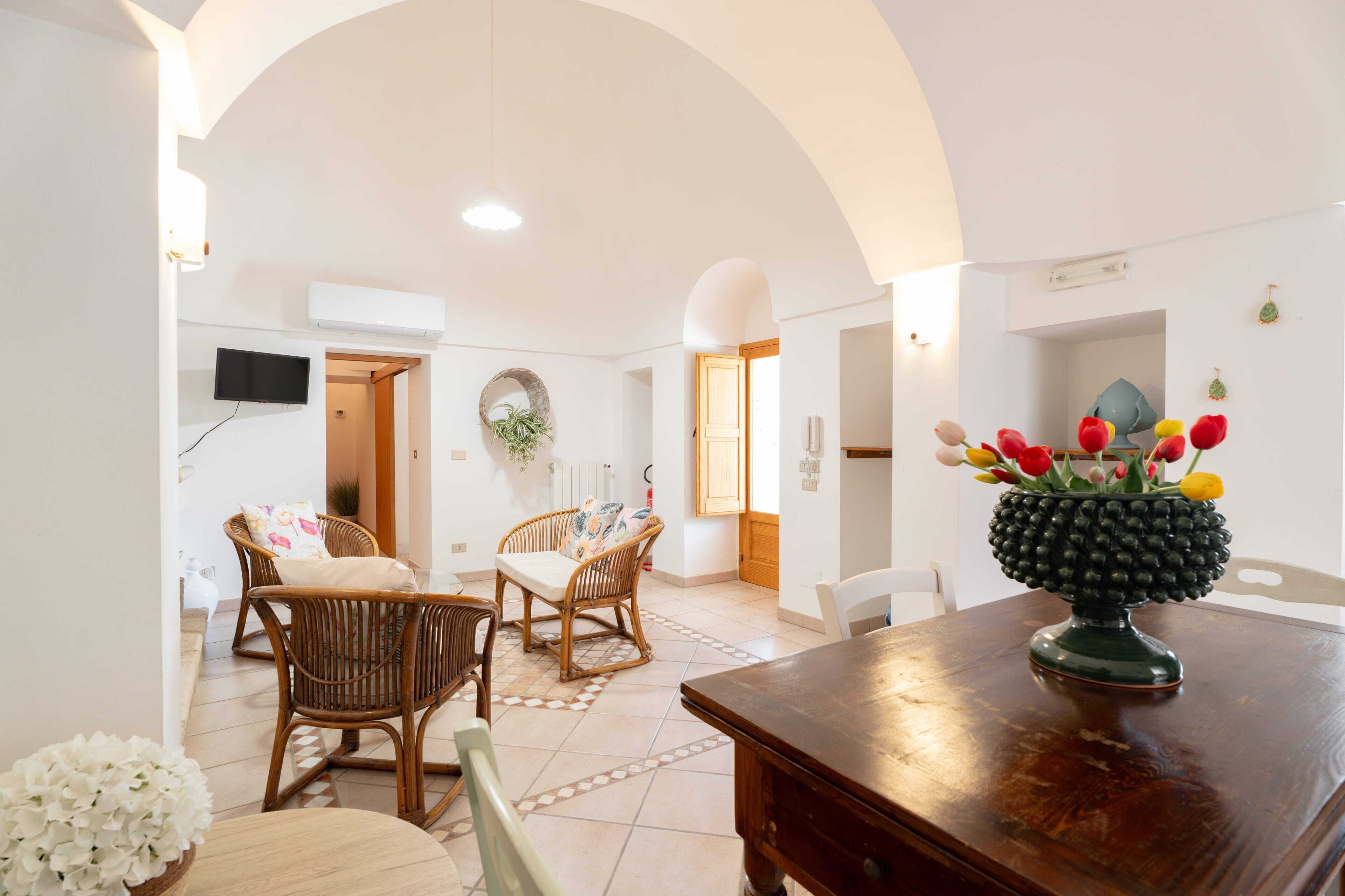 A spacious living area with a dining table, wicker chairs, and a decorative vase with flowers.