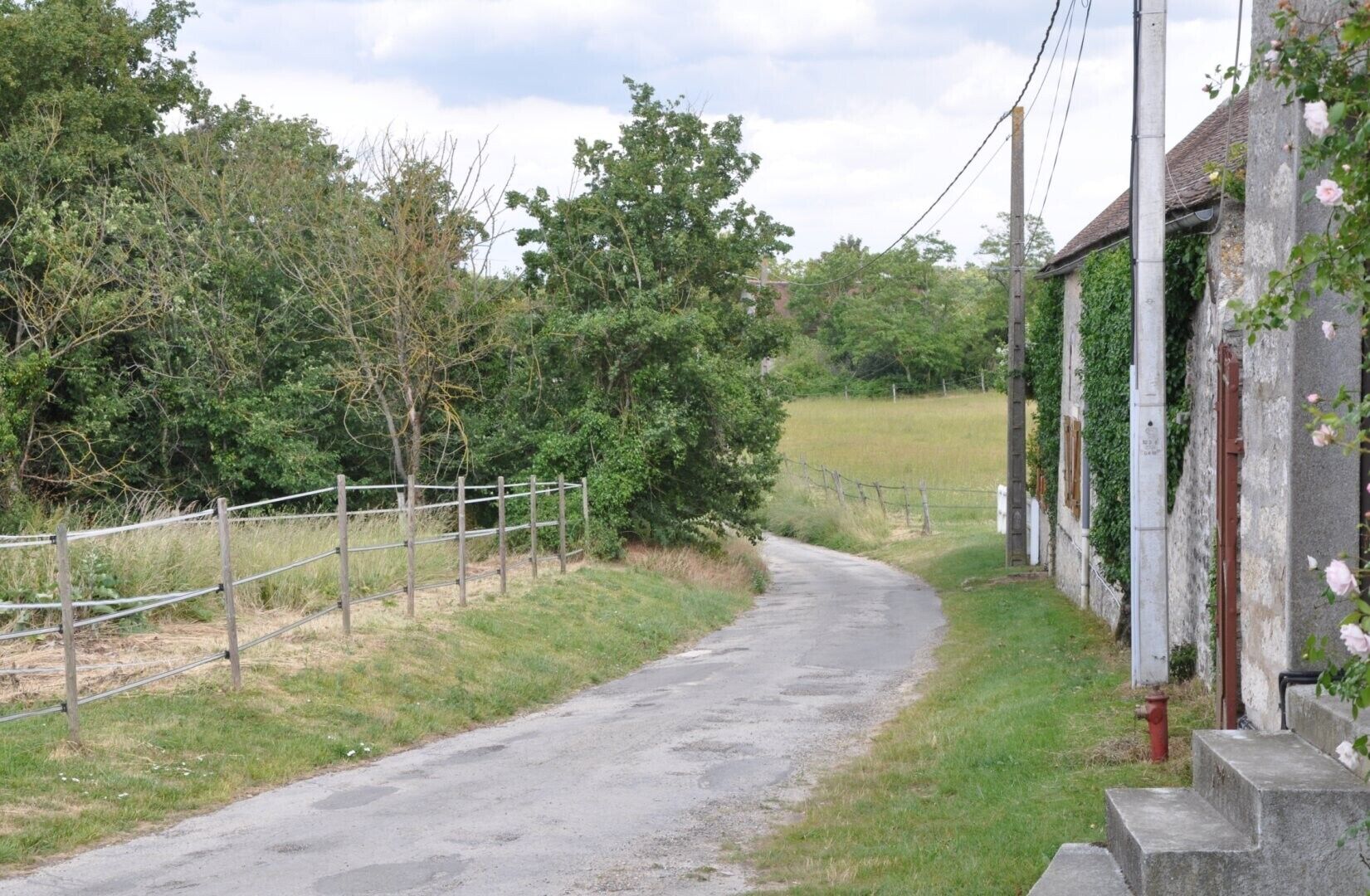 A rural road curving through a field with a fence on one side and a building with a garden on the other.