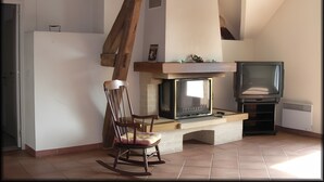 Een kamer met een open haard, een televisie, een schommelstoel en zichtbare houten balken.