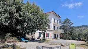 Un edificio de dos pisos con balcón, rodeado de árboles y un área de grava con mobiliario exterior.