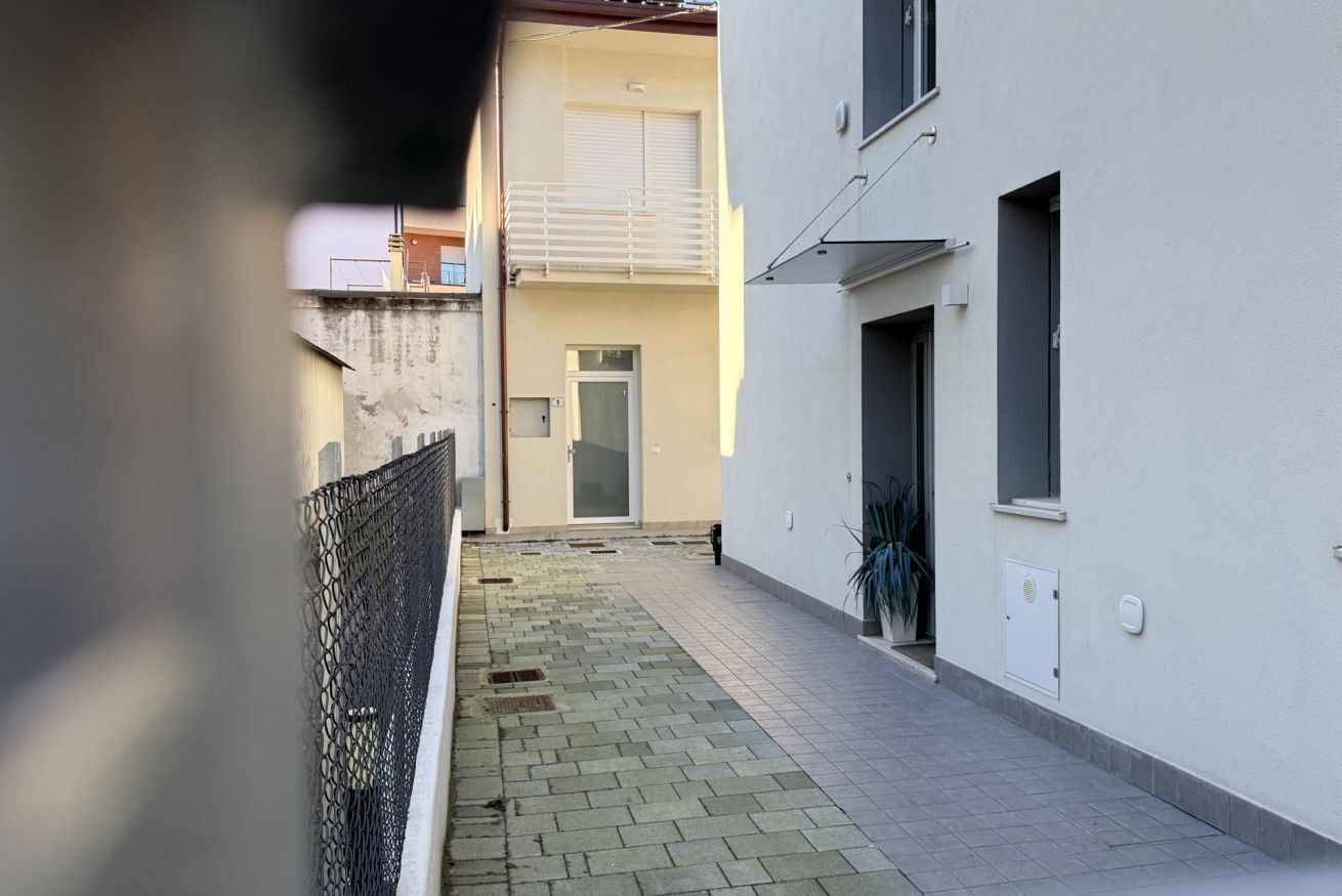 Uma viela estreita com um edifício branco à direita, uma porta e uma pequena janela.