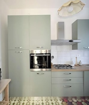 Cozinha com forno embutido, fogão a gás e micro-ondas. Há armários com puxadores e uma bancada com chaleira e máquina de café.