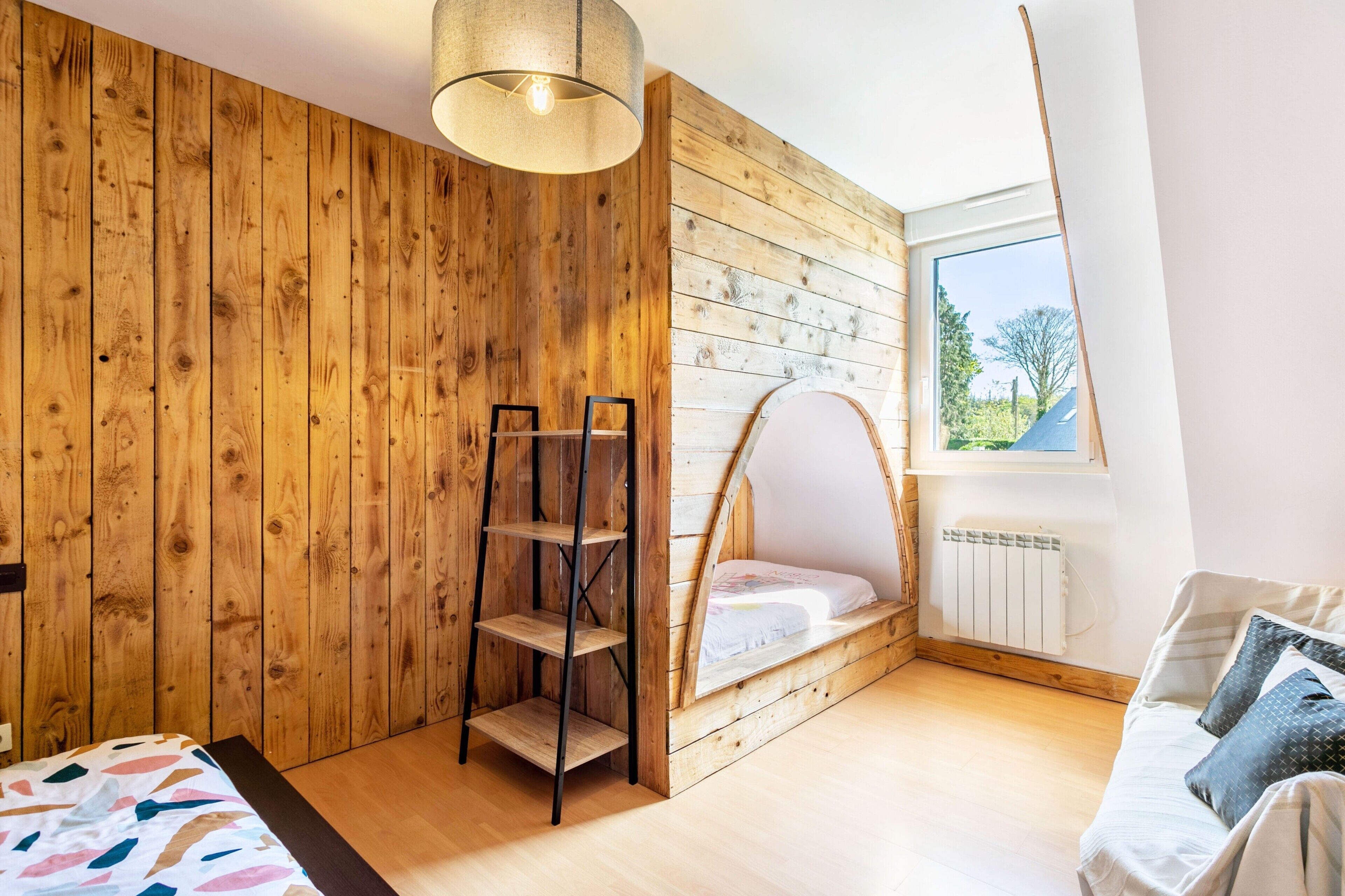 A bedroom with a wooden wall, a loft bed, a ladder, a sofa, and a window with a view.