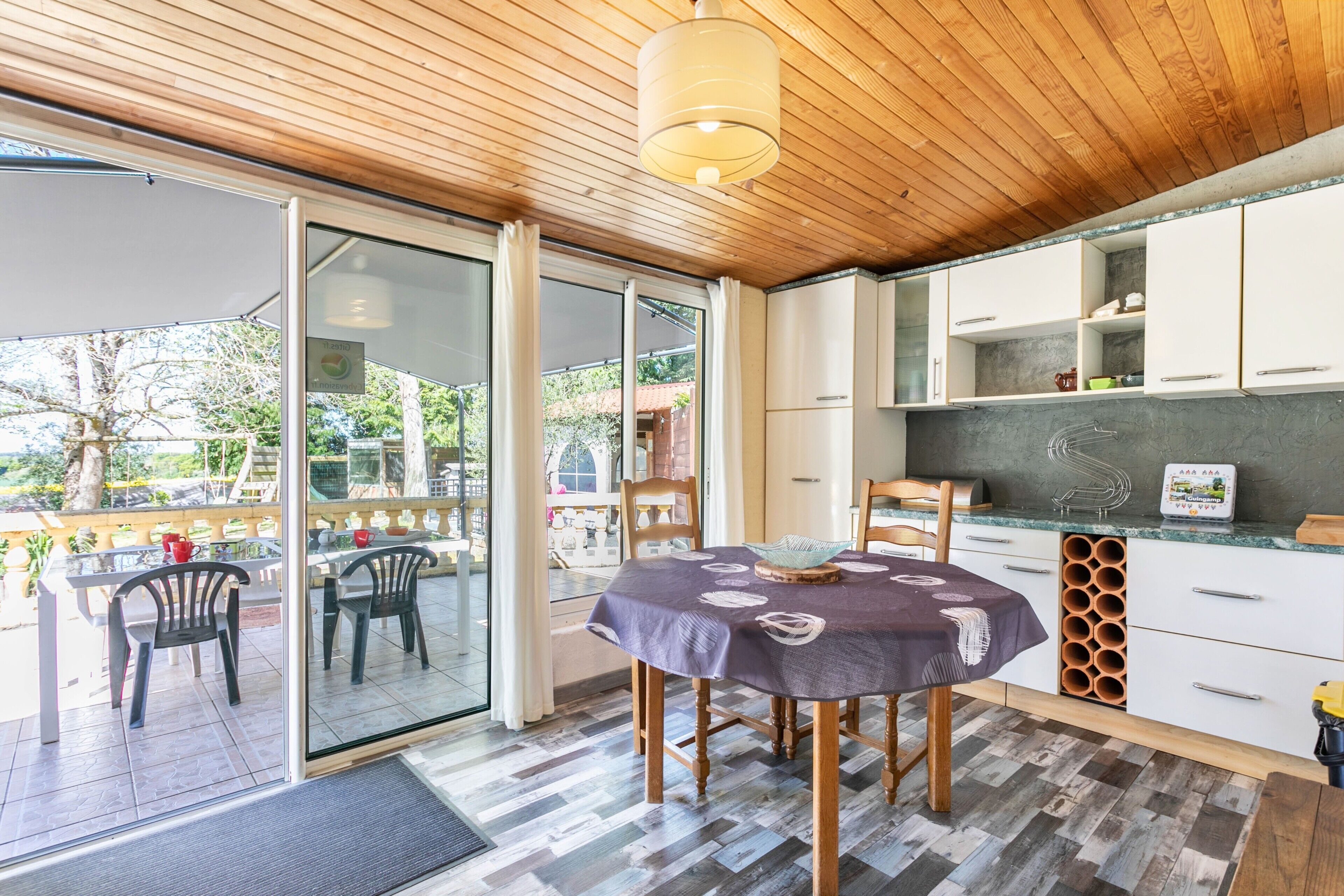 A modern kitchen with a dining area, wooden ceiling, and large windows.