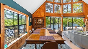 A large dining area with a long table, wooden walls, and large windows overlooking a lake. - Riverfront Retreat with Dock, Kayaks and Views (Shelby)