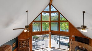 A room with a sloped ceiling, large windows, a ceiling fan, and a view of an outdoor deck. - Riverfront Retreat with Dock, Kayaks and Views (Shelby)