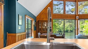 A kitchen with a stainless steel sink, wooden cabinetry, and a view of a lake through large windows. - Riverfront Retreat with Dock, Kayaks and Views (Shelby)