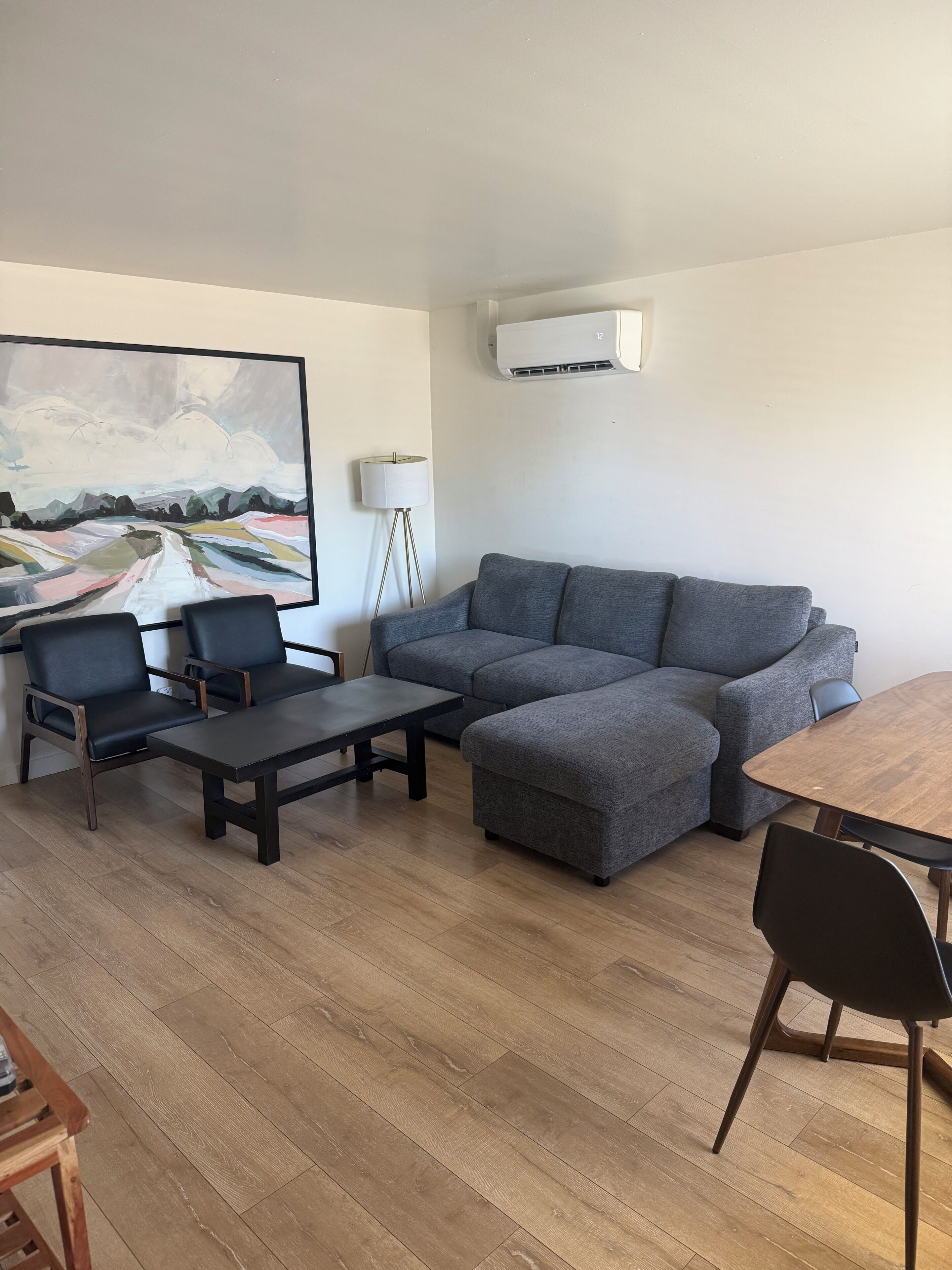 A living room with a sectional sofa, two armchairs, a coffee table, a dining table with chairs, a floor lamp, and a large abstract painting on the wall.