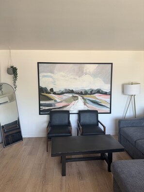 Un salon avec une grande peinture abstraite, deux fauteuils noirs, une table basse sombre, un canapé gris, un lampadaire et une plante suspendue.