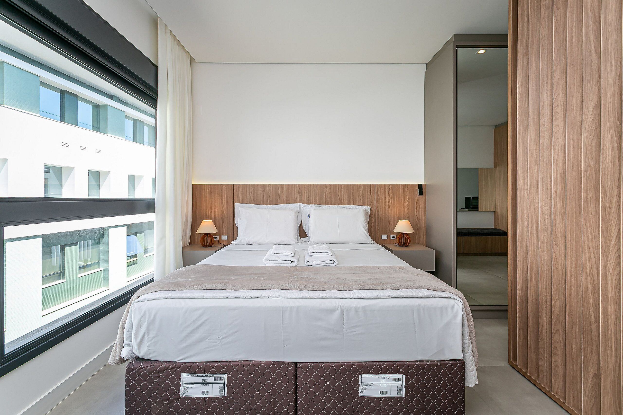 Une chambre d’hôtel moderne avec un grand lit, deux lampes de chevet, une vue par la fenêtre et une armoire en bois.