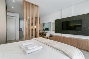 A modern hotel room with a bed, a TV mounted on the wall, a desk with a chair, and a wooden partition.