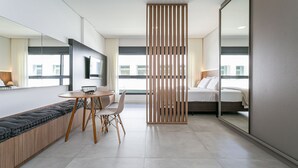 Una habitación de hotel moderna con sofá, mesa de comedor, sillas, cama, un espejo grande y vistas a la ciudad.