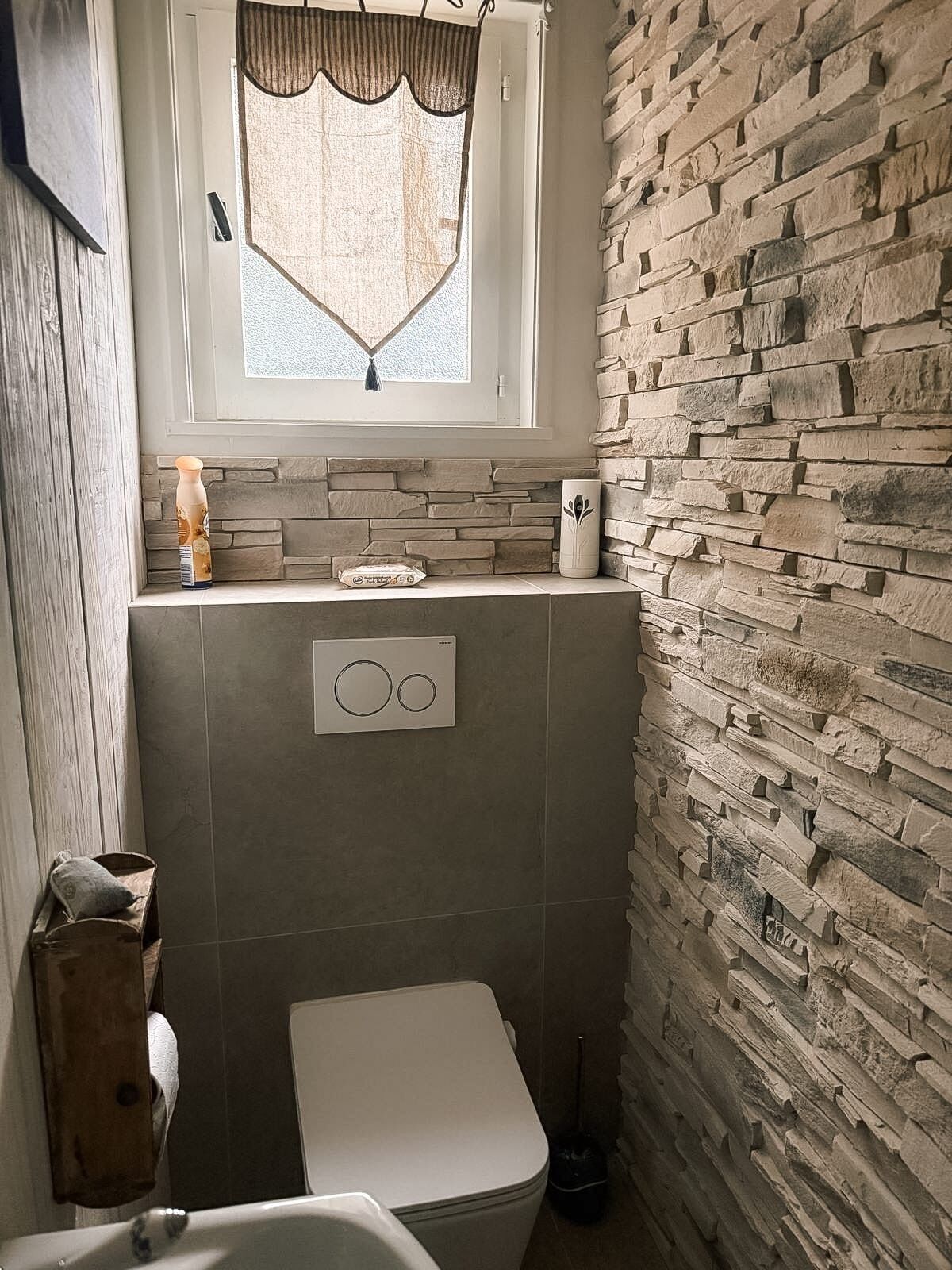 Een badkamer met een stenen muur, een raam met gordijn, een toilet en een douchehoek.
