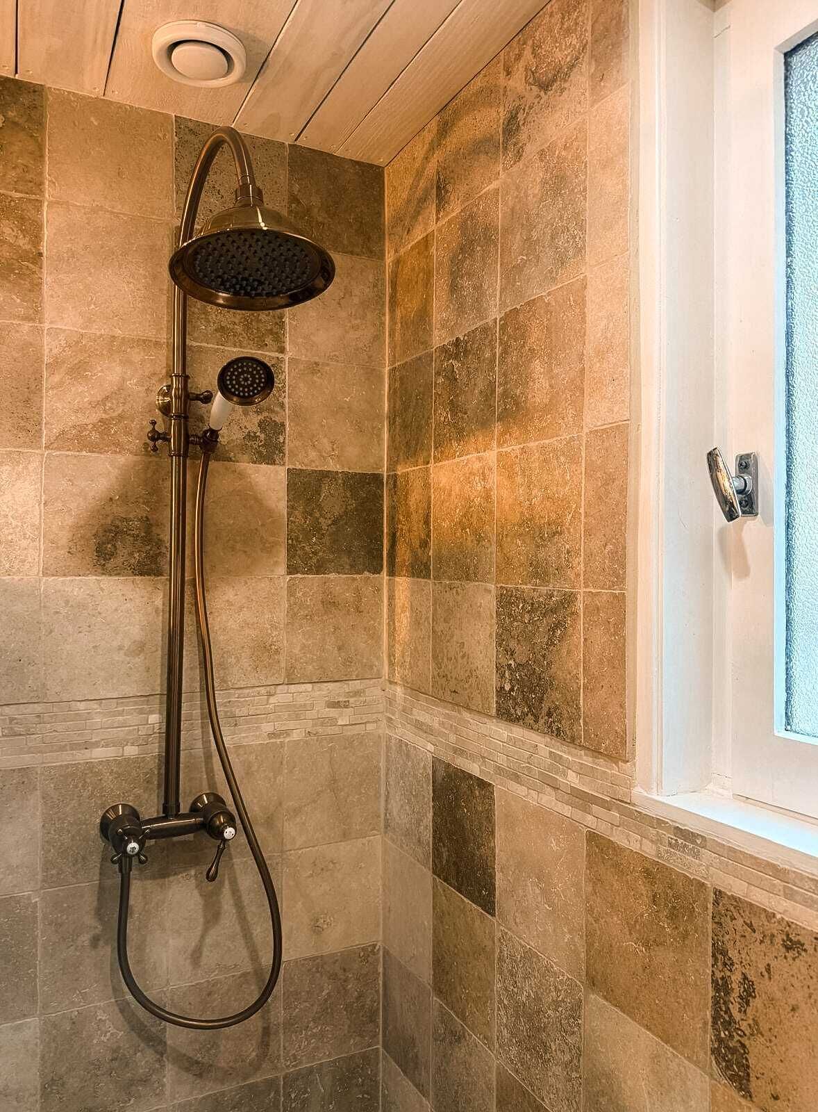A shower with a handheld showerhead and a wall-mounted showerhead, set against a tiled wall with a window to the right.