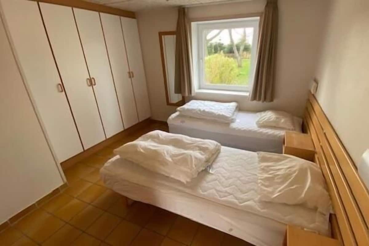 A bedroom with two beds, a wardrobe, and a window with a view of greenery.