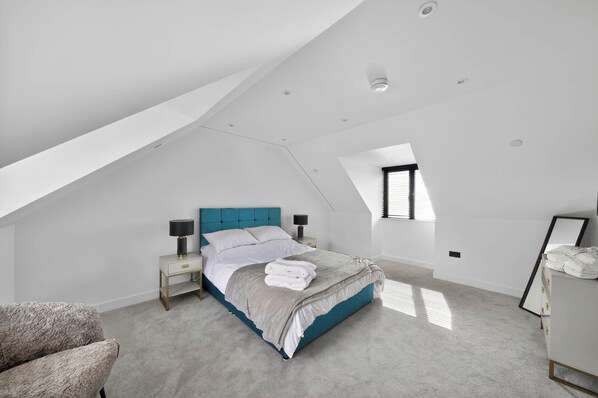 A bedroom with a sloped ceiling, a bed with a blue headboard, a bedside table with a lamp, and a window with blinds. - Heddfan (Carmarthen)