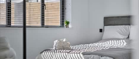 Una cama bien hecha con una colcha a rayas, una planta pequeña en maceta sobre el alféizar de la ventana y una ventana con persianas.