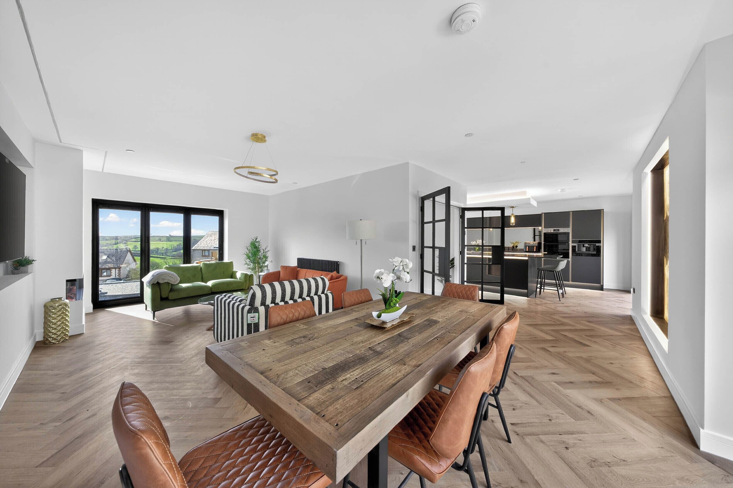 Una sala moderna con una mesa de comedor de madera, sillas de cuero, un sofá verde y vistas a través de amplios ventanales.