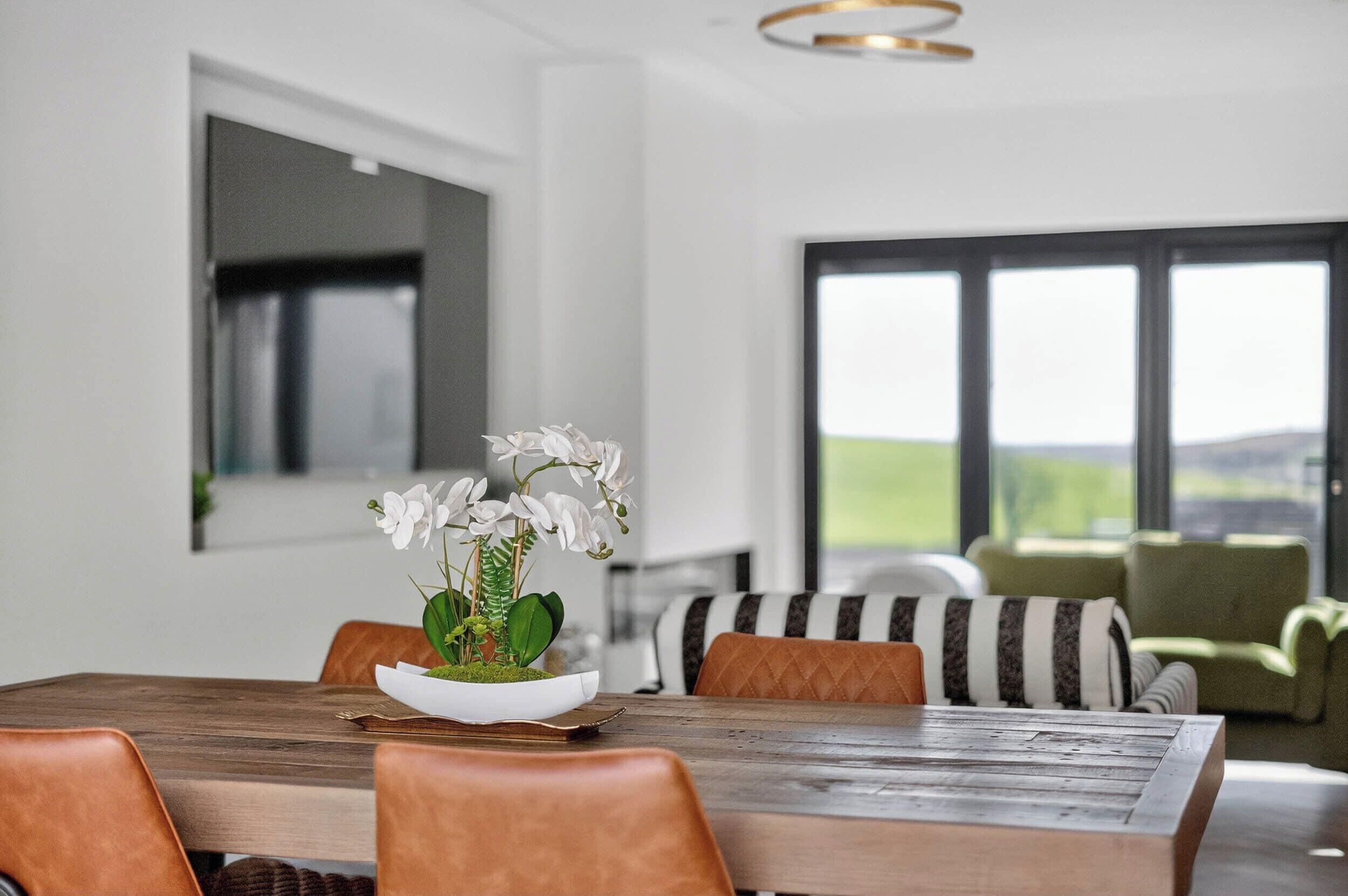 A dining area with a wooden table, brown leather chairs, a white orchid centerpiece, and a view of a green landscape through large windows.