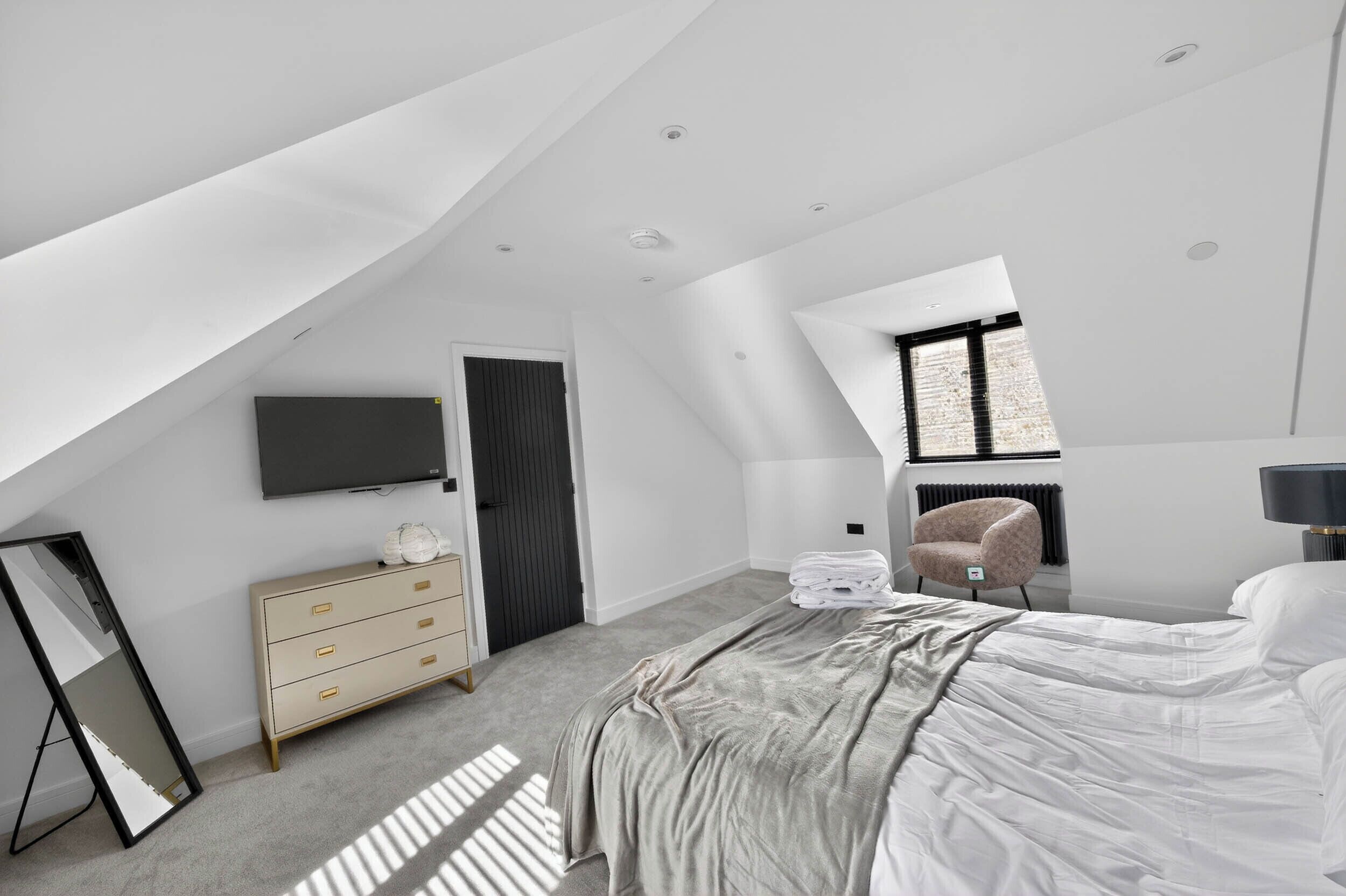 Un dormitorio moderno con techo inclinado, una cama, un aparador, un televisor y una silla.