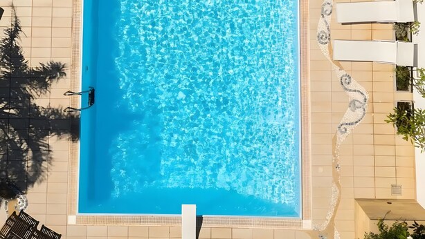 Ein rechteckiger Pool mit klarem, blauem Wasser, umgeben von Fliesenböden und weißen Liegestühlen.