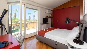 Ein modernes Hotelzimmer mit Bett, Schreibtisch mit Laptop, Fernseher und Balkon mit Blick auf Palmen.
