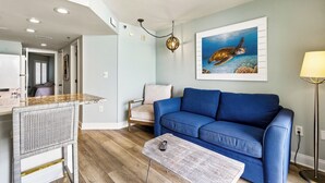 Un salón con un sofá azul, un cuadro de una tortuga marina, una silla de mimbre y una zona de cocina con barra.
