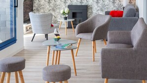 Un salon moderne avec des fauteuils à revêtement gris, une petite table ronde sur laquelle repose un livre, et un fauteuil blanc agrémenté d’un coussin rouge.