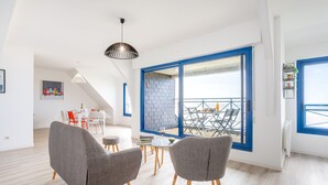 Un salon moderne avec des fauteuils à revêtement gris, un coin repas avec des chaises blanches, une porte coulissante à cadre bleu donnant sur un balcon et un luminaire suspendu.