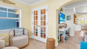 A room with a large window, a door with glass panels, a chair with a cushion, a small table, and a basket. - Barefoot Beach Resort E206 - Liquid Obsession (Indian Shores)