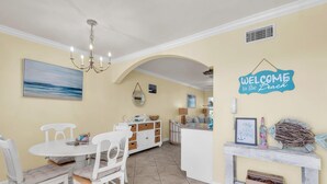 A dining area with a white table and chairs, a chandelier, and a 'Welcome to the Beach' sign. - Barefoot Beach Resort E206 - Liquid Obsession (Indian Shores)