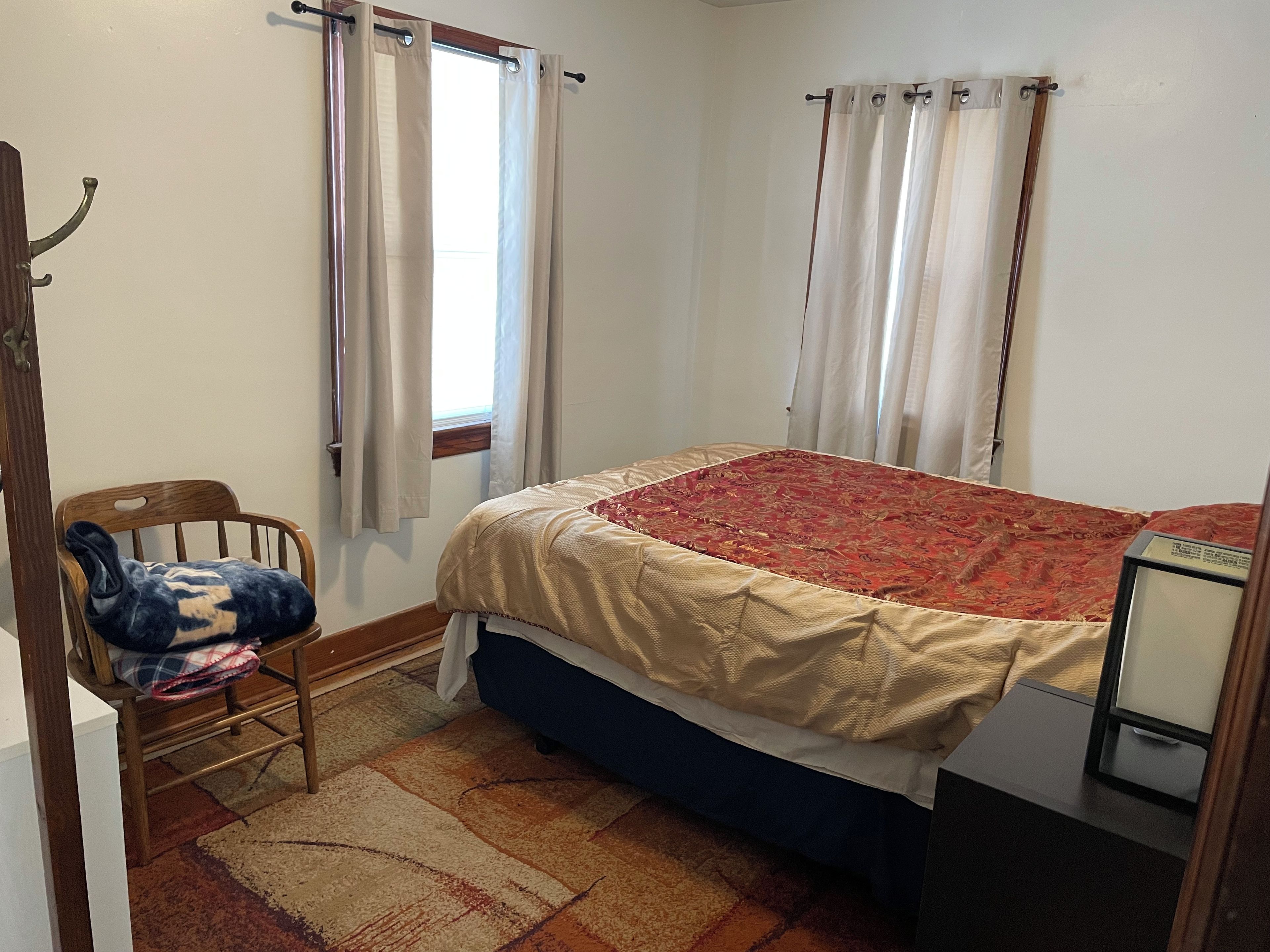 Un dormitorio con cama, una silla, un ventanal con cortinas y una mesita.