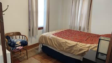 Un dormitorio con cama, una silla, un ventanal con cortinas y una mesita.
