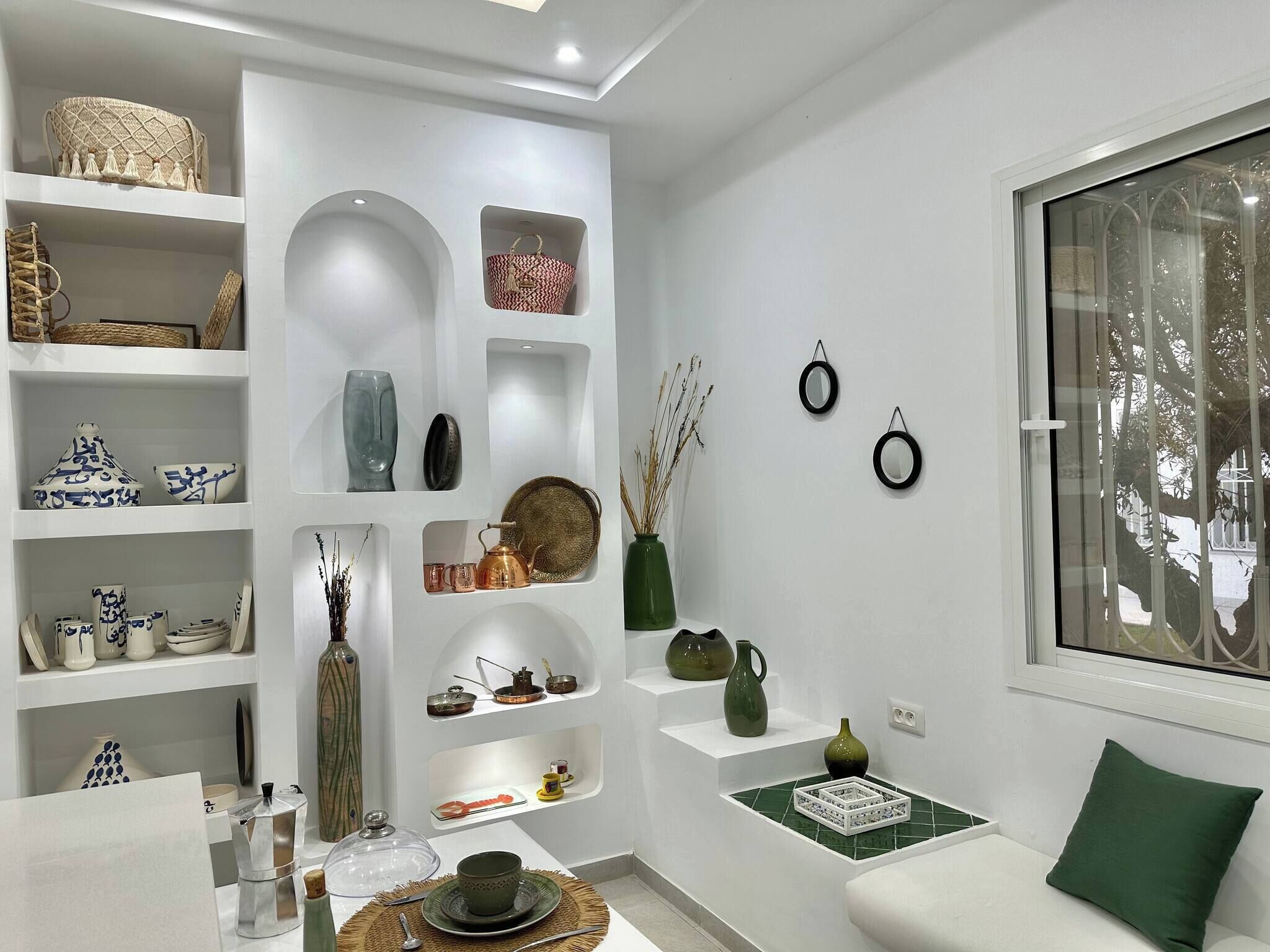 Un intérieur moderne avec des murs blancs, des étagères ornées de divers objets décoratifs, un coin salon avec des coussins verts et une fenêtre donnant sur des arbres.